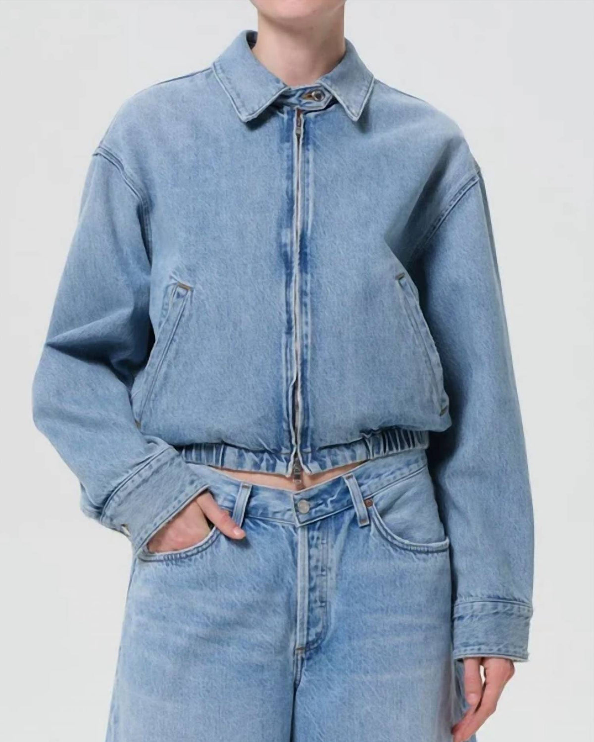 A model wearing a cropped, light-wash blue denim jacket with a front zipper and collar, paired with matching wide-leg denim jeans, against a plain light gray studio background.